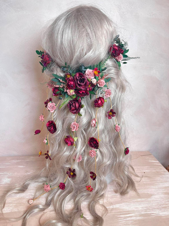 Long wavy silver-gray wig with a floral hair accessory featuring burgundy, pink, and green flowers, displayed on a mannequin head against a soft neutral background.