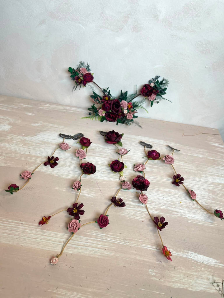 A floral garland or decorative branch arrangement with burgundy and pink faux flowers, green leaves, and small bird-shaped embellishments, laid out on a light beige surface against a textured off-white wall.