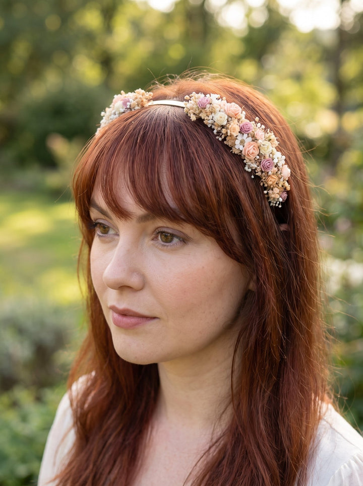 A woman with red hair wearing a delicate floral headband with small pastel flowers, photographed outdoors against a blurred green garden background.