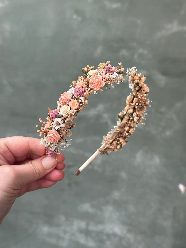 A hand holding a floral headband decorated with pink and peach roses, small white flowers, and beige botanical elements, set against a textured gray background.