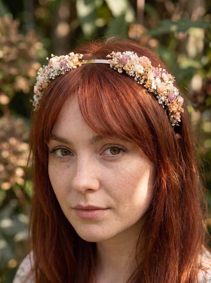 A close-up portrait of a woman with red hair wearing a delicate floral crown made of small cream, pink, and peach blossoms, set against a soft-focus outdoor background of green foliage.
