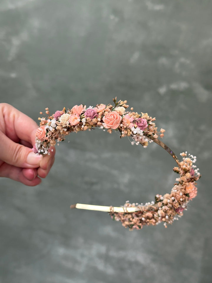 A hand holding a delicate floral headband decorated with pink and peach roses, small white flowers, and green leaves, set against a textured gray background.