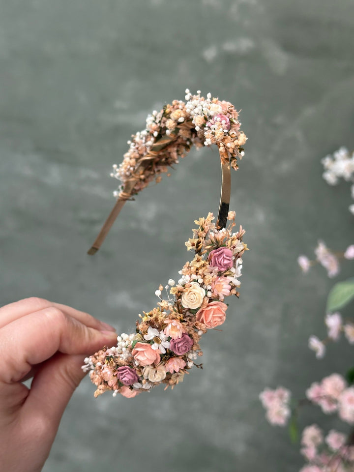 A hand holds a curved floral headband decorated with small pink and peach roses, cream blossoms, and baby's breath against a soft gray background with blurred pink flowers.