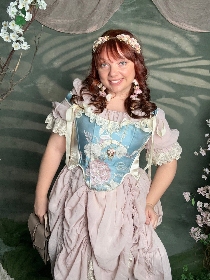 A woman wearing a vintage-style blue and pink dress with lace ruffles, a floral headpiece, and matching earrings, posing in front of a green leafy backdrop with cherry blossom decorations.