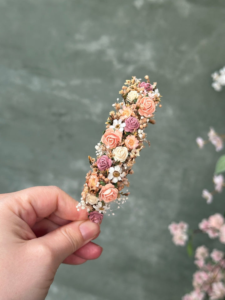 A hand holding a delicate flower headband with small peach, pink, cream, and mauve dried roses and baby's breath against a soft gray-green blurred background.