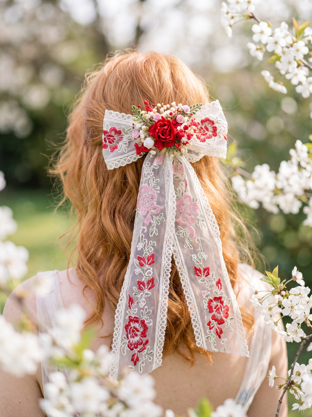 A red-haired woman viewed from behind outdoors, wearing a decorative hair bow accessory with white lace, pink floral embroidery, and a red rose at the center, surrounded by soft white blossoms.