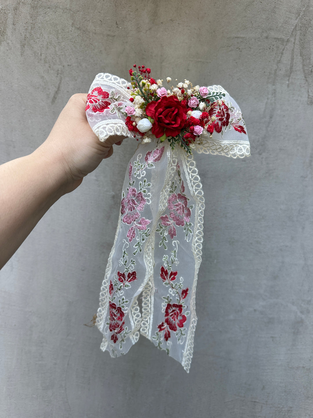 A hand holding a decorative hair bow accessory featuring red and pink embroidered lace ribbons, white lace trim, and a floral center with red roses, pink filler flowers, and small white pearls, set against a textured gray wall.