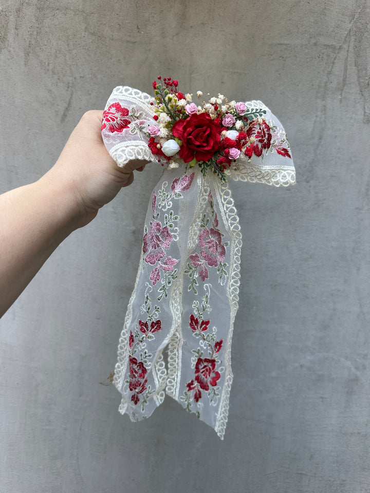 A hand holding a decorative hair bow accessory featuring red and pink embroidered lace ribbons, white lace trim, and a floral center with red roses, pink filler flowers, and small white pearls, set against a textured gray wall.