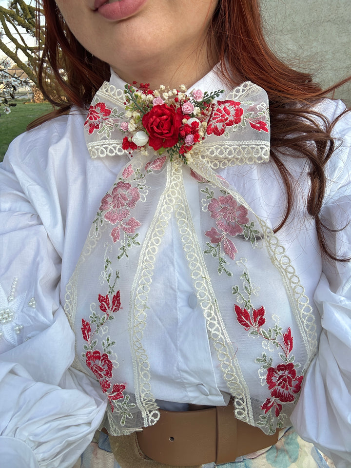 A close-up of a person wearing a white blouse with a large decorative bow at the collar. The bow is made of cream lace and features red and pink floral embroidery, green leaves, and a central red rose embellishment with small pearl-like beads.