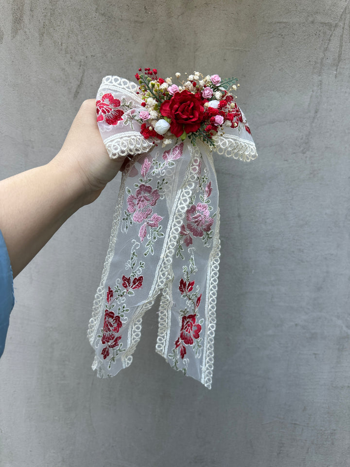 A hand holding a decorative bridal veil or hair bow accessory with a red floral bouquet at the top and long sheer white lace ribbons printed with pink and red flowers, set against a gray concrete wall.