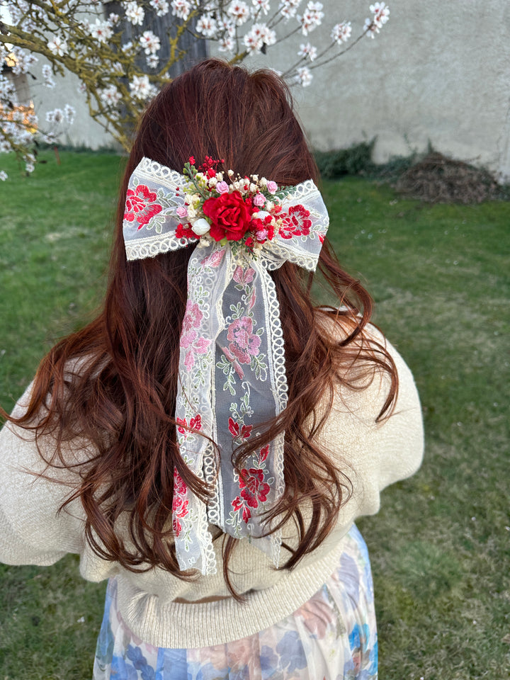 A woman viewed from behind wearing a large decorative hair bow in her brown hair. The bow features red and pink floral lace trim, white lace, and a central red rose and floral cluster.