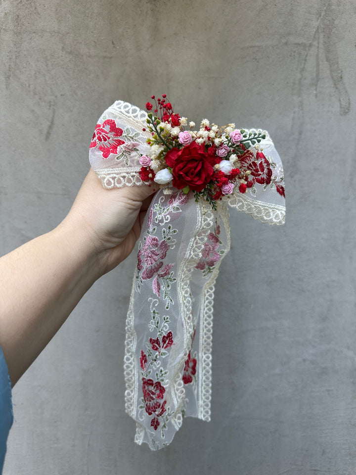 A hand holding a decorative hair accessory featuring a white lace bow with red floral embroidery, a red rose, and small pink and white flowers against a textured gray wall.
