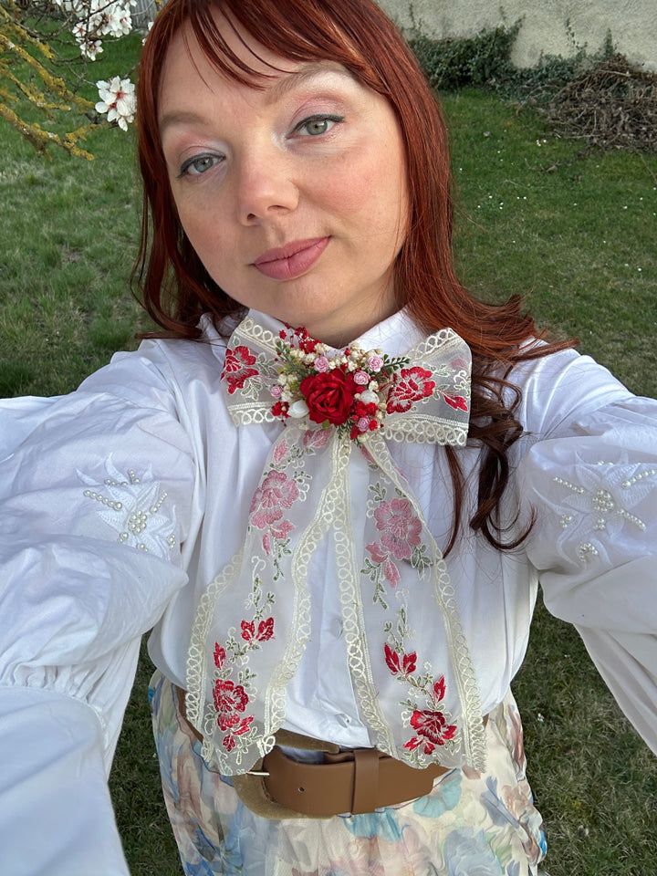 A woman with red hair takes a selfie outdoors wearing a white long-sleeve blouse with floral embroidery, a large cream lace bow decorated with red roses, and a floral skirt.