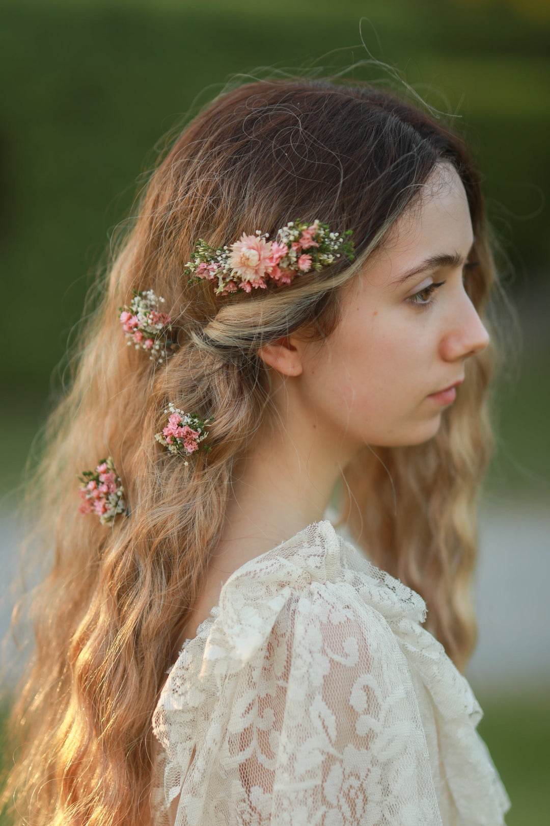 A woman in profile wearing a cream lace top and a floral hair accessory with pink flowers and green leaves, photographed outdoors against a blurred green background.