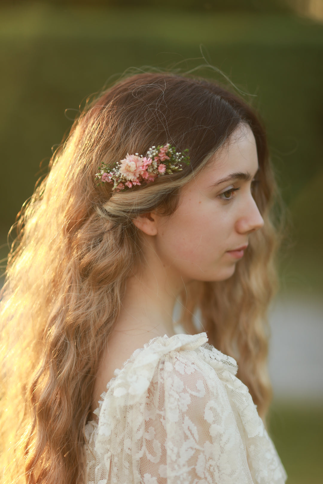 A woman in profile wearing a delicate pink and white floral hair accessory, with long wavy light brown hair and a cream lace top, set against a soft blurred green background.