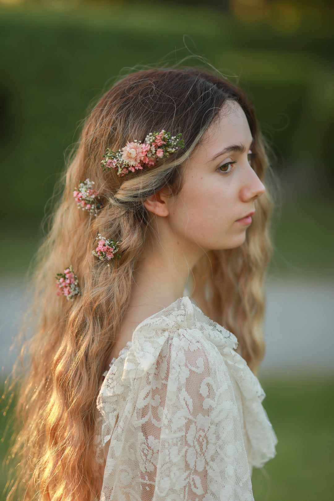 A woman in profile wearing a cream lace top and a floral hair accessory with pink flowers and green leaves, photographed outdoors against a blurred green background.