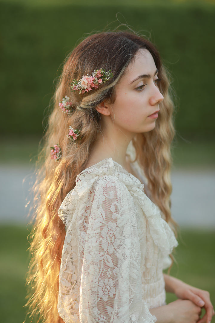 A woman in profile wearing a cream lace blouse and a floral hair accessory with pink flowers and green leaves, photographed outdoors against a blurred green background.