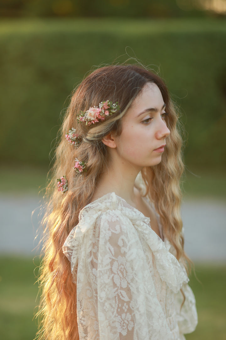 A woman with long wavy light brown hair wearing a floral crown and a cream lace blouse, photographed outdoors against a soft green blurred background.