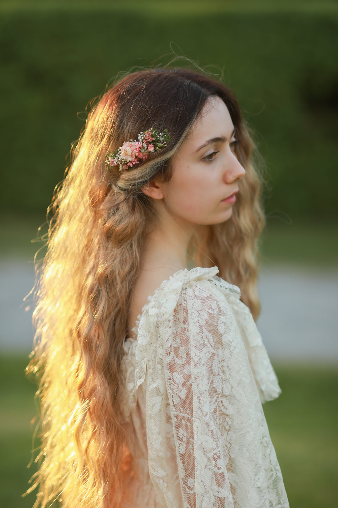 A young woman with long, wavy light brown hair wearing a delicate floral hair accessory and a cream lace top, photographed outdoors in warm golden hour light against a blurred green background.