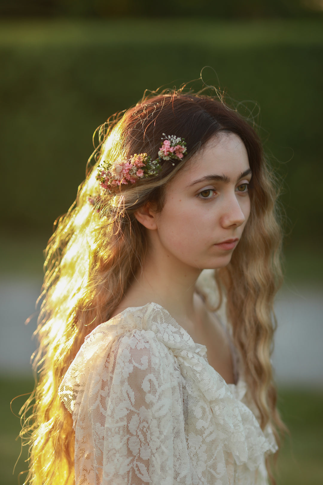 A woman with long wavy light brown hair wearing a delicate pink and green floral headpiece, dressed in a cream lace top, set against a soft-focus green outdoor background.