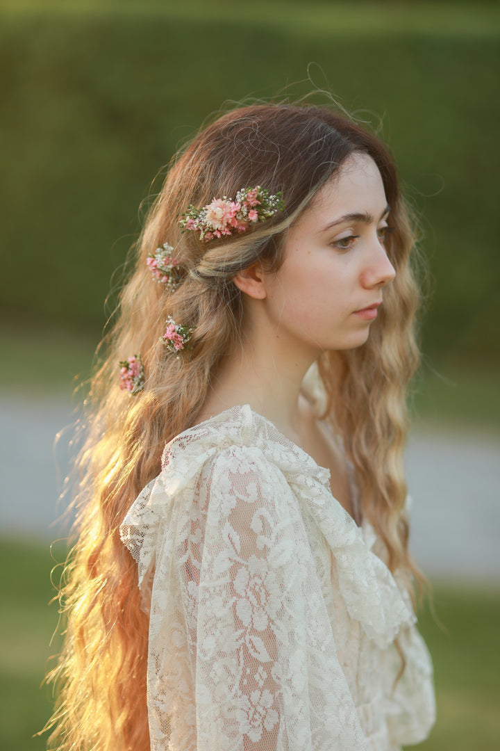 A woman in profile wearing a cream lace blouse and delicate pink floral hair accessories, photographed outdoors with soft warm light and a blurred green background.