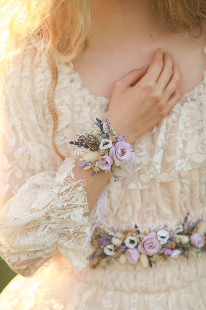 Close-up of a person wearing a floral wrist corsage in purple and cream tones with a lace dress.