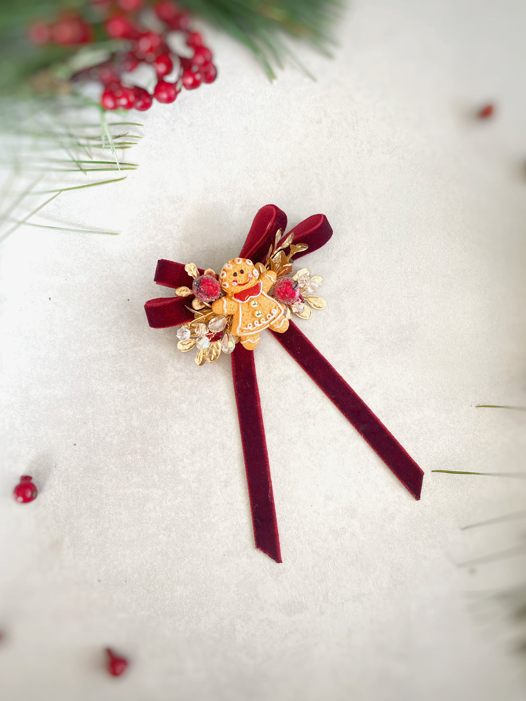 A festive Christmas ornament brooch featuring a gold gingerbread man figure, red velvet ribbon tails, and red and pink gemstone accents, displayed on a textured off-white surface with holiday pine and berry decorations.