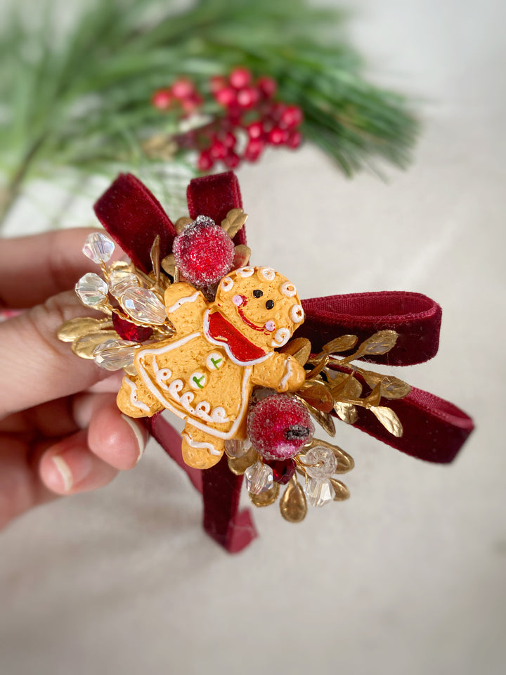 A hand holds a Christmas-themed hair bow accessory featuring a gingerbread cookie ornament, red velvet ribbon, gold leaf accents, and red berries against a soft neutral background with blurred pine branches.