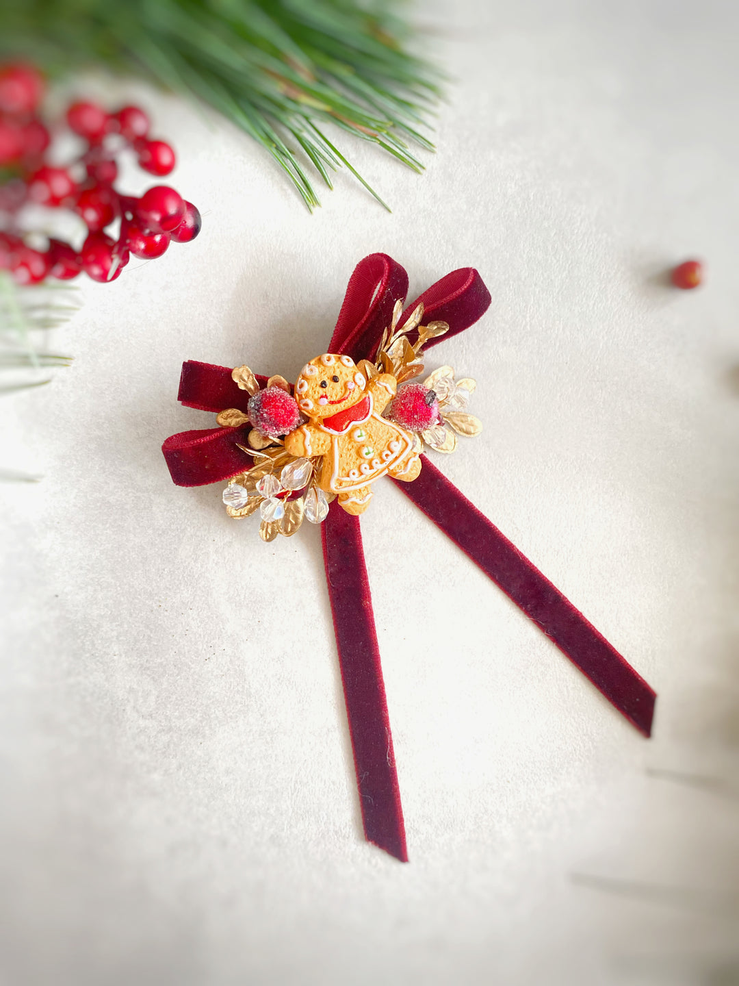 A festive Christmas-themed hair accessory featuring a gold-plated angel figure, red velvet ribbon tails, and red faux berries, displayed on a textured light gray background with holiday pine needles and red berries as props.