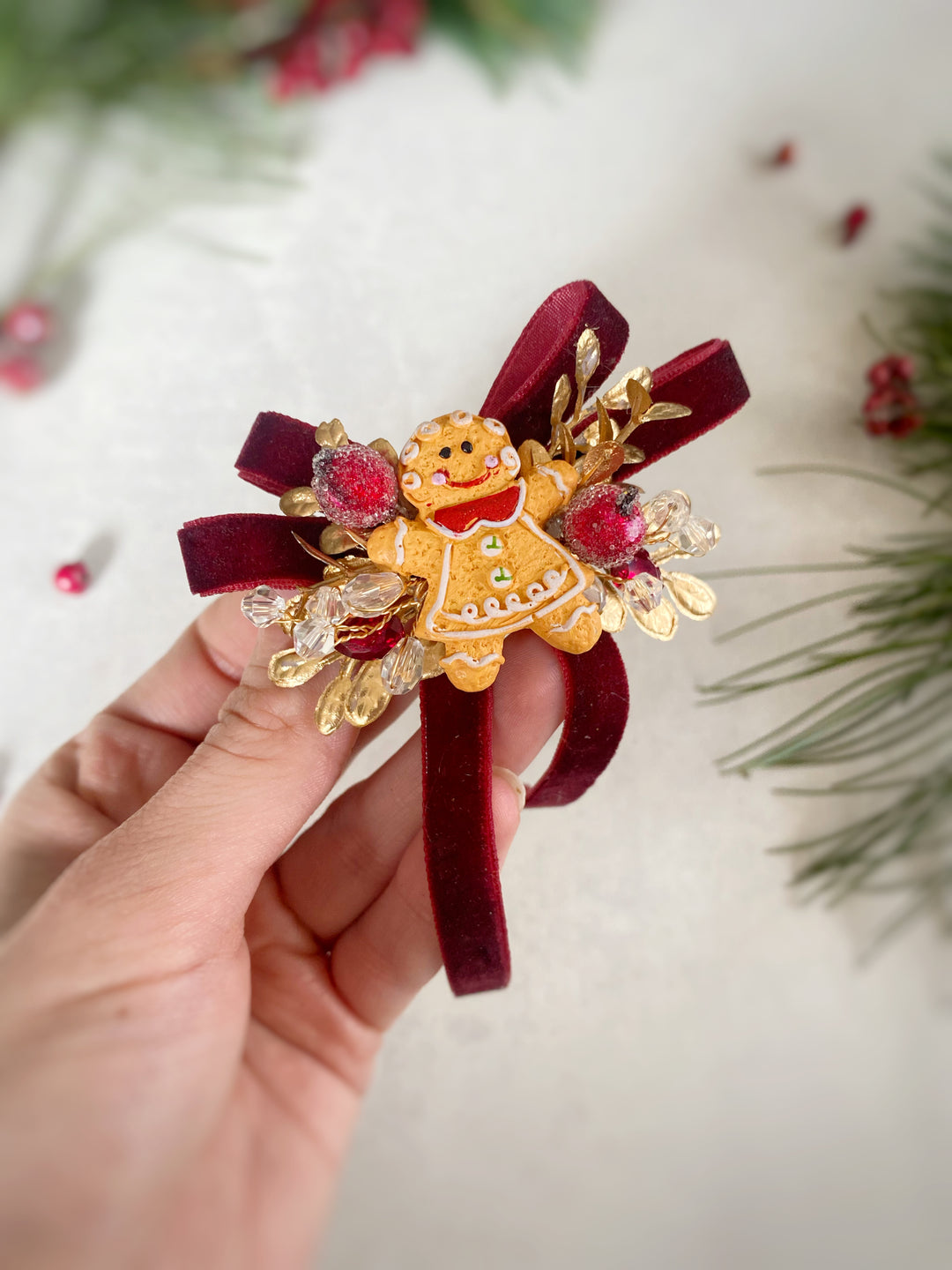 A hand holding a festive Christmas-themed hair claw clip. The clip features a gold gingerbread man figure, deep burgundy velvet ribbon bows, and red and gold faux berries, set against a softly blurred holiday background.