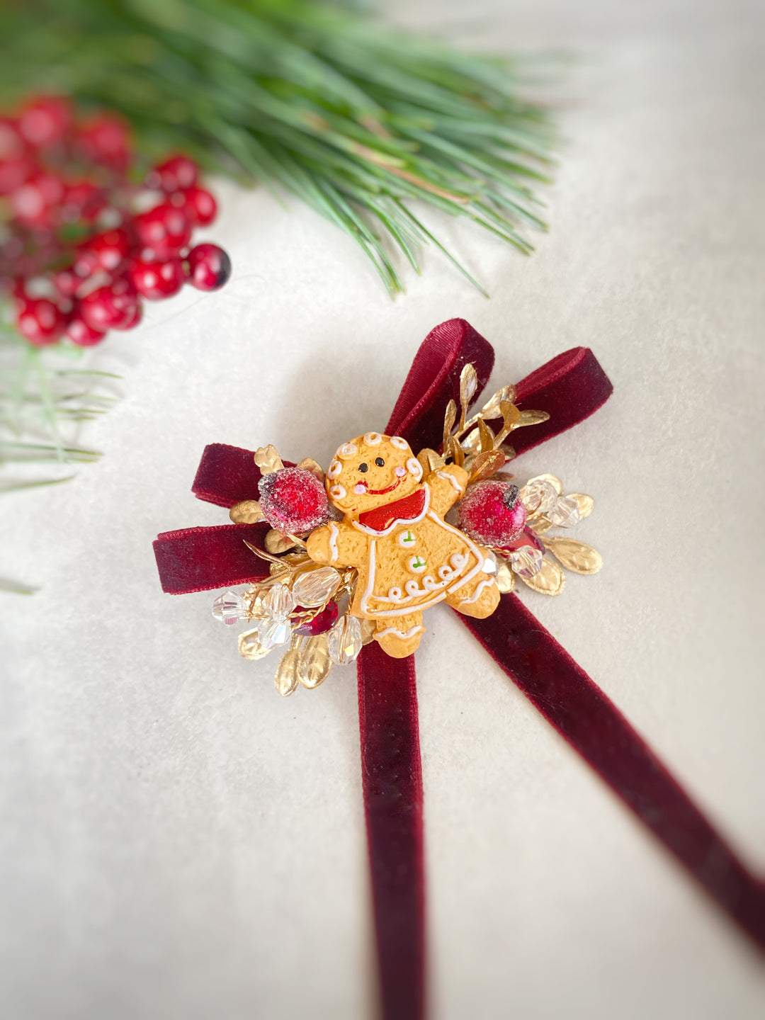 A Christmas-themed hair bow accessory featuring a gold gingerbread person figure, red berries, and a burgundy velvet ribbon, styled with pine branches and red berries on a light beige background.