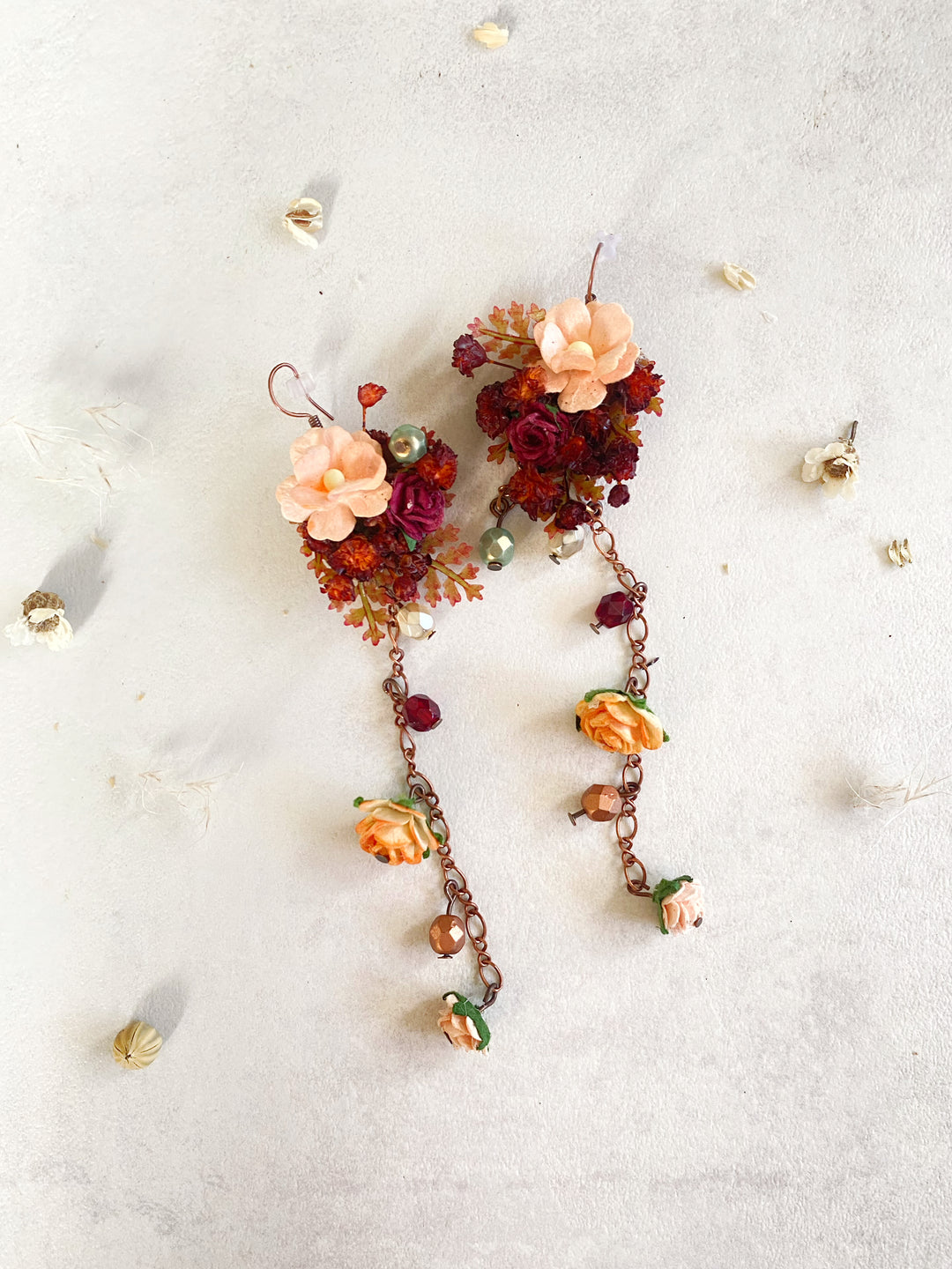 A pair of handmade dangle earrings featuring peach and orange flowers, deep red dried floral clusters, and copper-toned chains with small beads, laid flat on a textured off-white surface with scattered dried flower petals.