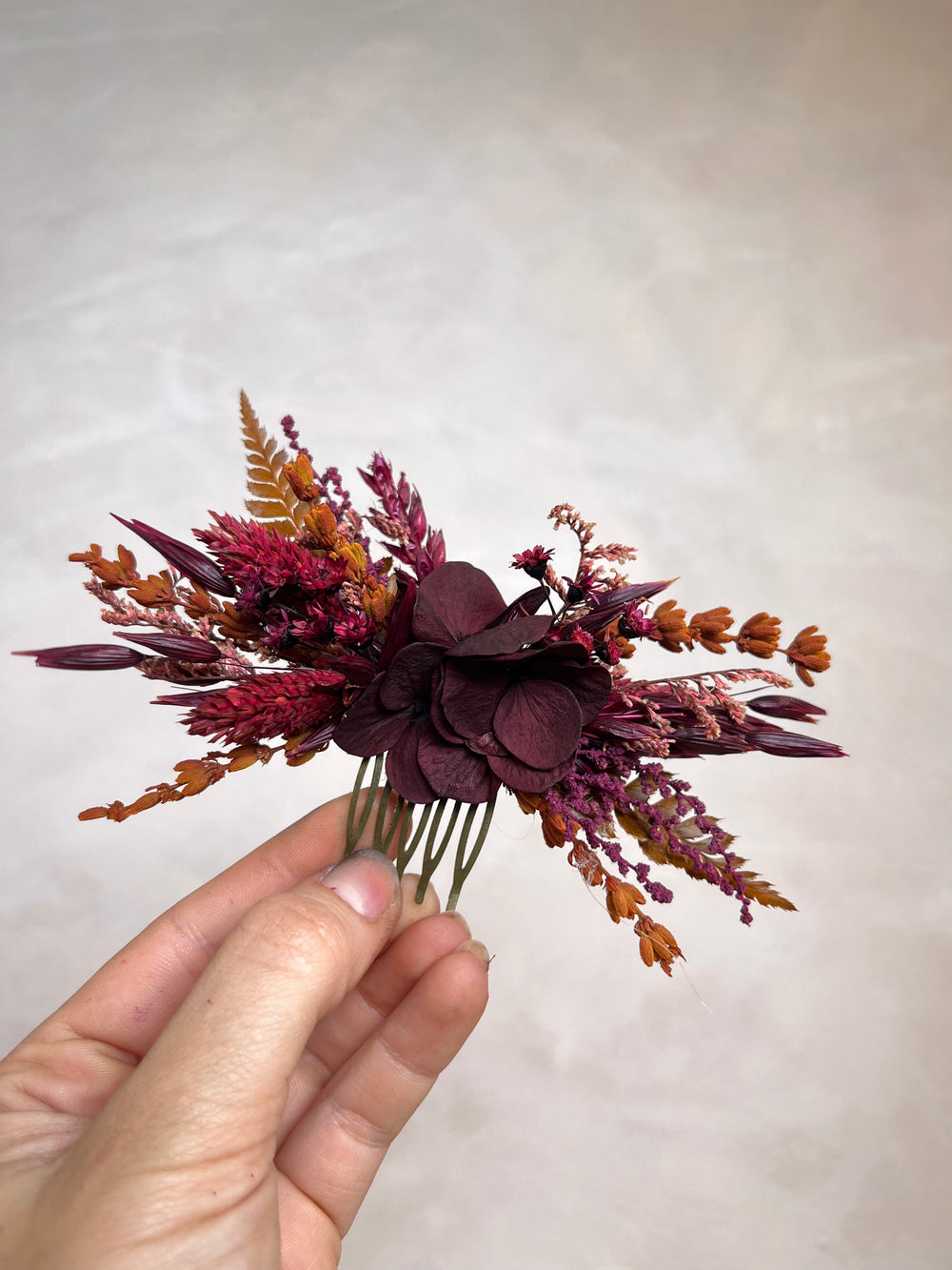 A hand holding a dried flower hair comb accessory featuring deep burgundy hydrangea petals, rust-colored ornamental grasses, against a neutral beige background.