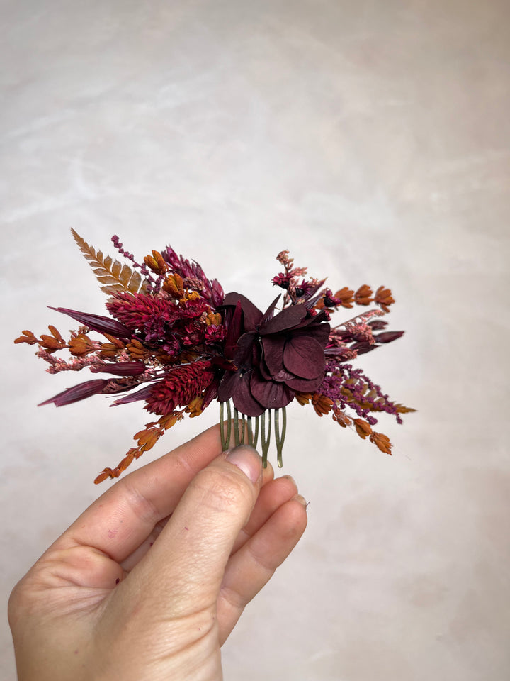 A hand holding a dried floral hair comb featuring burgundy, deep purple, and rust-orange botanical elements against a neutral beige background.