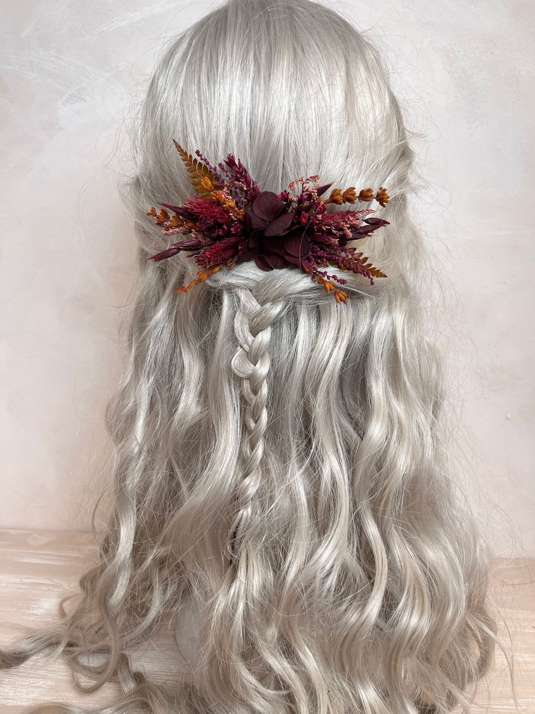 Long wavy silver-gray wig with a side braid, styled with a dark burgundy and orange dried floral hair accessory, shown against a soft beige background.
