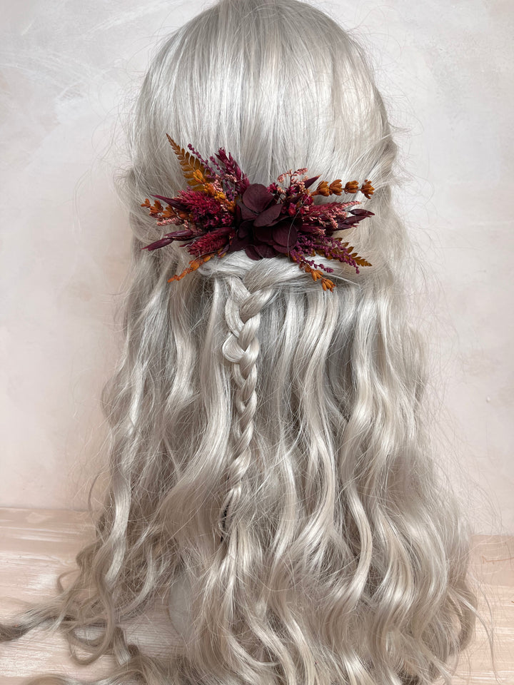 Long wavy silver-gray wig with a side braid, styled with a dark burgundy and orange dried floral hair accessory, shown against a soft beige background.