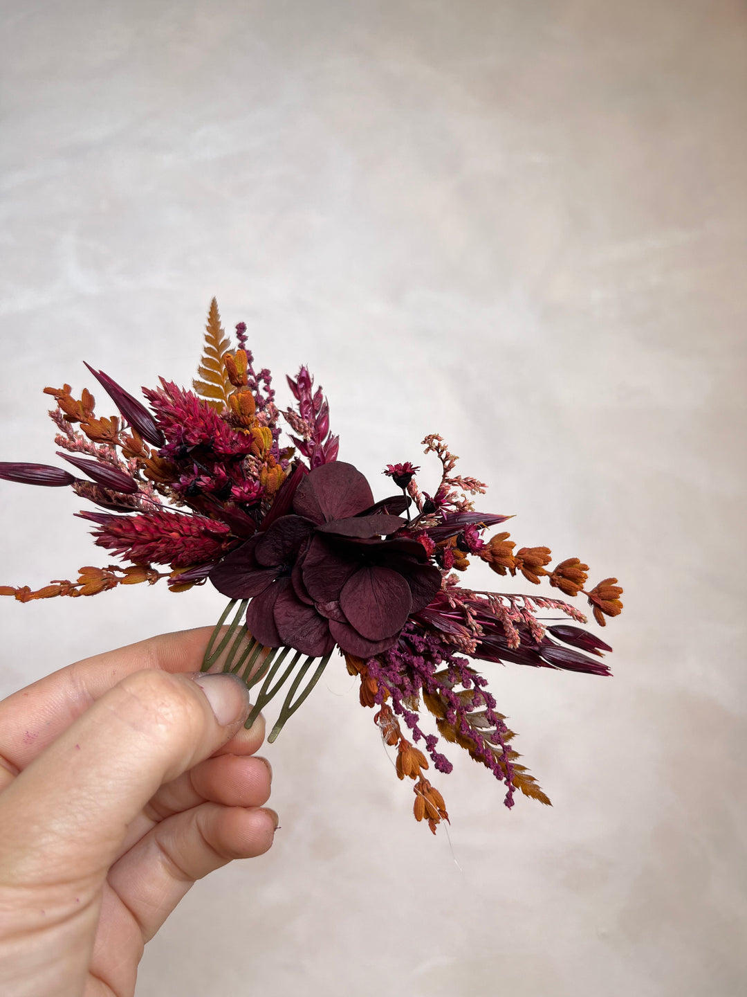 A hand holding a dried floral hair comb featuring burgundy, deep purple, and rust-orange botanical elements against a neutral beige background.