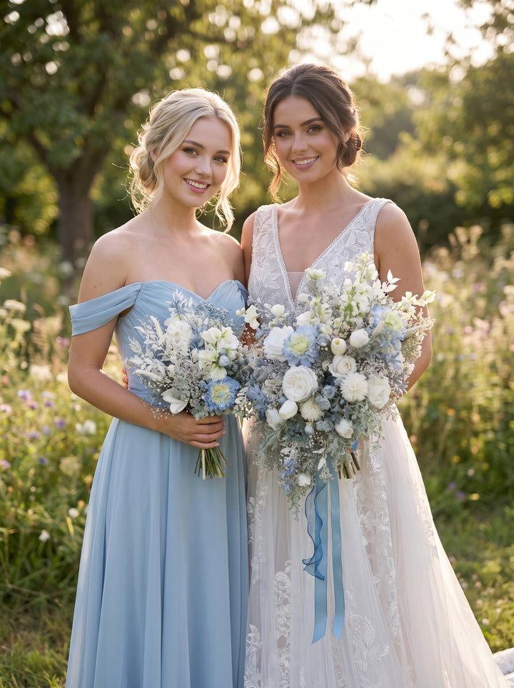 A bride and bridesmaid standing outdoors in a sunlit field. The bridesmaid wears an off-the-shoulder light blue gown and holds a bouquet of white and pale blue flowers, while the bride wears a sleeveless white lace wedding dress and holds a matching bouquet with blue ribbon accents.