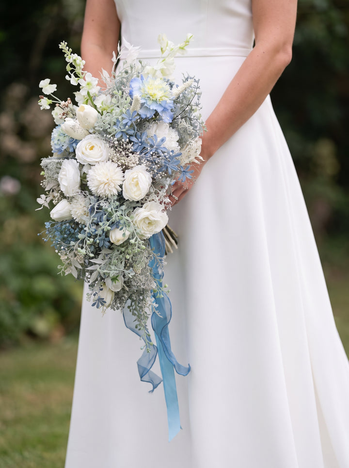 A bride in a white wedding gown holding a bridal bouquet of white and light blue flowers with a blue ribbon, photographed outdoors against a blurred green background.