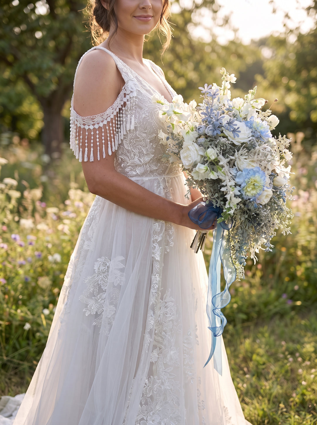 A woman outdoors in a white lace wedding dress with beaded fringe on the sleeves, holding a large bouquet of white and light blue flowers.