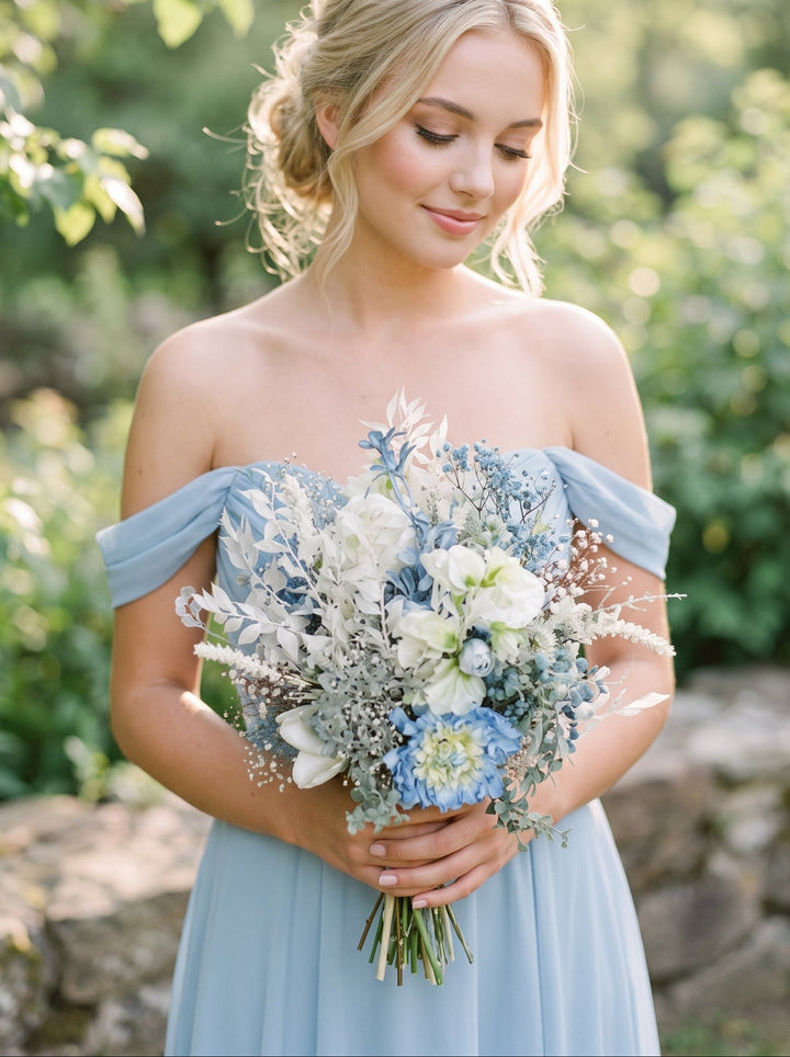 A blonde woman outdoors wearing an off-the-shoulder light blue dress and holding a bouquet of white and blue wildflowers, with a blurred green garden background.