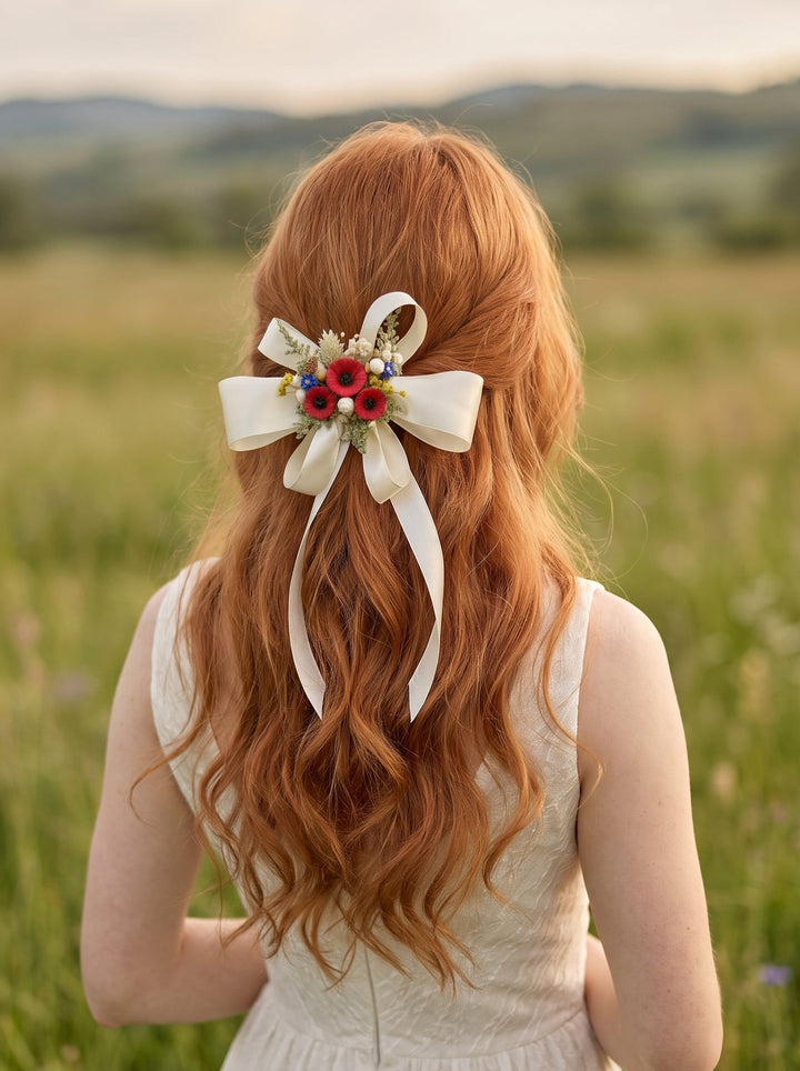 A woman with long wavy red hair seen from behind, wearing a white sleeveless dress and a white hair bow decorated with small red poppies, standing in a grassy field with hills in the background.