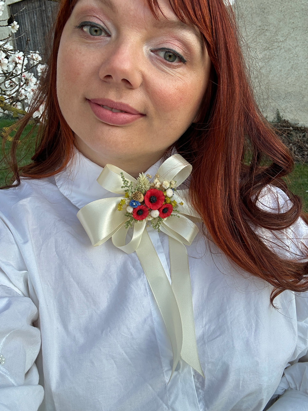 A close-up selfie of a woman with red hair wearing a white high-neck blouse and a decorative cream satin bow at the collar adorned with small red flowers and greenery.