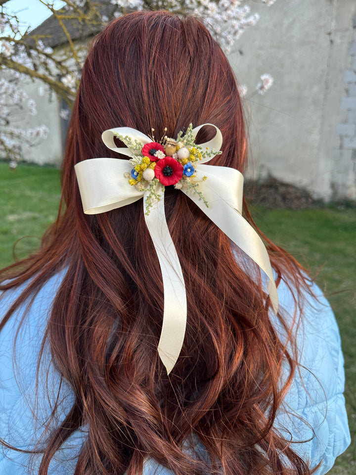 A long, wavy auburn hair accessory featuring a large cream satin bow and a colorful floral cluster with red, yellow, and blue flowers, shown worn on a woman's hair in an outdoor setting.