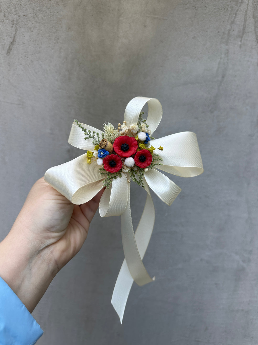 A hand holding a small decorative floral wristlet or corsage with a large cream ribbon bow, red poppy-style flowers, and small green and blue filler accents against a gray textured wall.