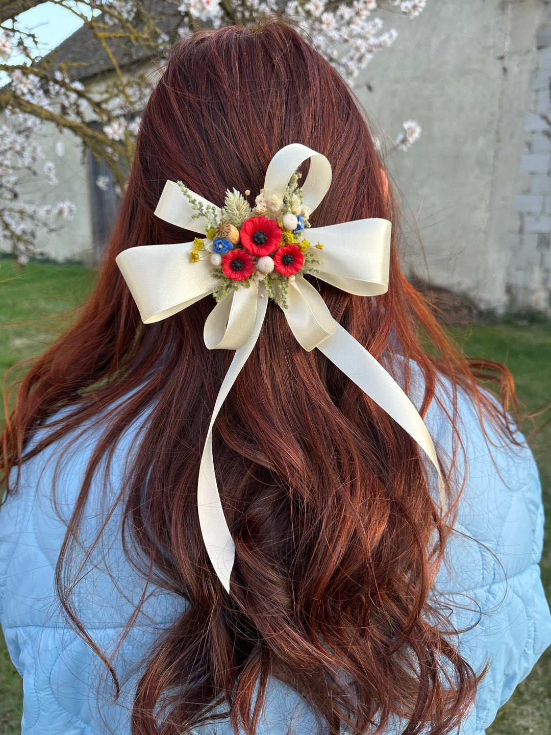 A long wavy auburn hair accessory featuring a large cream satin bow and a central floral cluster with red poppies, yellow flowers, and small blue details, shown pinned to the back of a woman's hair.