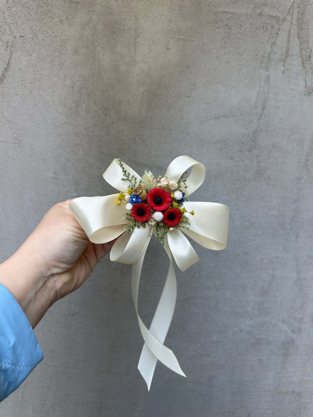 A hand wearing a light blue sleeve holds a small decorative hair bow  against a textured gray concrete wall. The bow features a large cream satin bow with long tails, a central cluster of red artificial flowers, tiny white pearls, and dried green foliage.