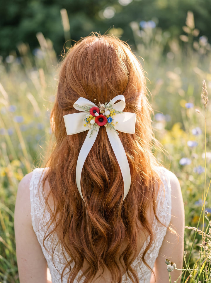 A woman with long wavy red hair seen from behind, wearing a white lace top and holding a large white ribbon bow with small red, yellow, and blue flowers in a sunlit grassy field.