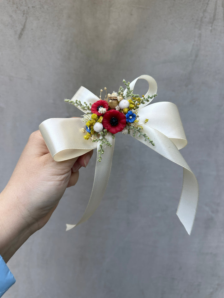 A hand holding a cream satin ribbon bow decorated with red, blue, and yellow dried flowers and green filler stems, set against a textured gray concrete wall.