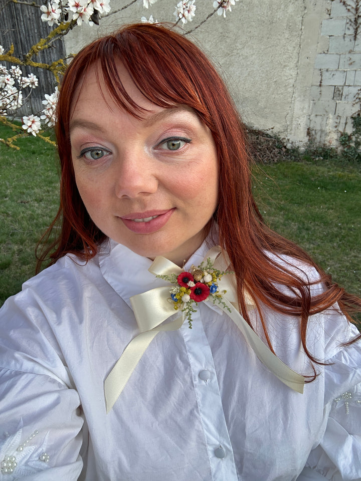 A woman with red hair wearing a white high-neck blouse with a large cream satin bow and a small floral boutonniere, photographed outdoors in front of a stone wall and flowering branches.